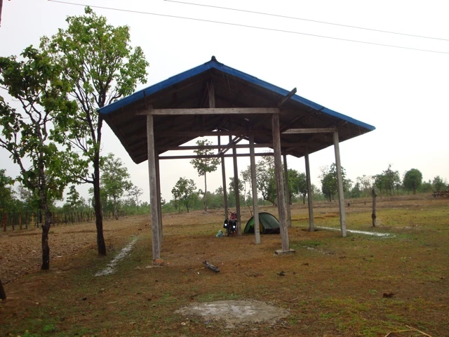 Offene Holzhütte mit blauem Dach auf karger Wiese, daneben ein grünes Zelt und ein Motorrad