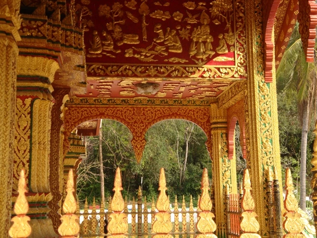 Detailreiche Nahaufnahme eines traditionellen laotischen Tempels mit goldverzierten Säulen, ornamentalen Bögen und Ziergiebeln vor Tropenwald