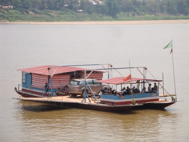 Großes Holzboot mit Überdachung transportiert Passagiere auf einem breiten, braunen Fluss mit grüner Uferlandschaft
