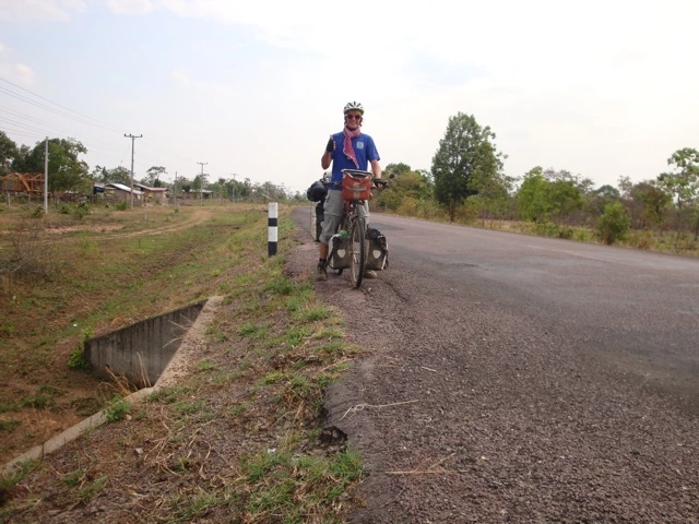 Radfahrer mit Gepäcktaschen auf einer asphaltierten Landstraße, links ein Straßengraben, Bäume und Felder im Hintergrund