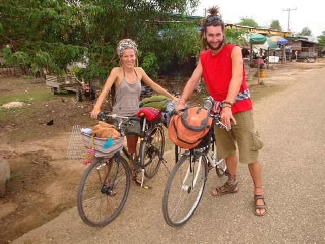 Zwei Fahrradtouristen mit beladenen Rädern auf unbefestigter Straße, umgeben von Bäumen und einfacher Bebauung
