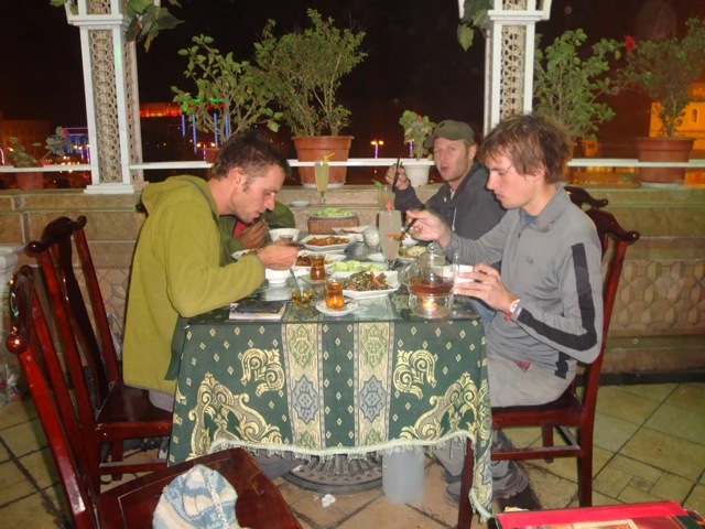 Drei junge Männer sitzen an einem gedeckten Tisch mit gemusterter Tischdecke, essen und trinken Tee