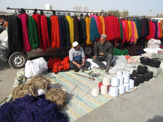 Marktstand mit hängenden mehrfarbigen Textilien, zwei Händler sitzen auf Decke neben Garn und Stoffen