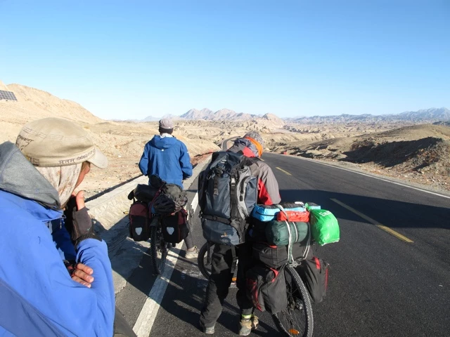 Mehrere Fahrradfahrer mit beladenen Rädern fahren auf einsamer Asphaltstraße durch felsige Wüstenlandschaft mit Bergkulisse