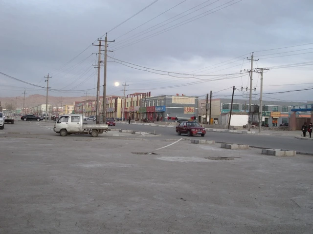 Verlassene Straßenkreuzung mit Strommasten, Berglandschaft im Hintergrund, graue Bebauung und parkende Fahrzeuge