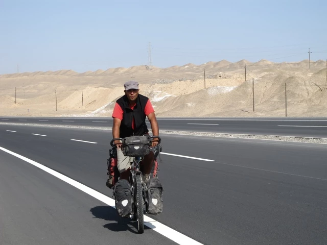 Fahrradfahrer mit Gepäcktaschen auf einer leeren Straße durch eine trockene Wüstenlandschaft mit Hügeln im Hintergrund