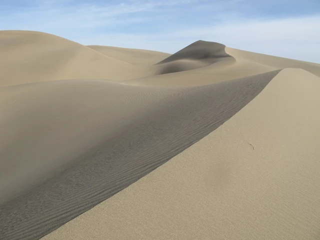 Weite Sandlandschaft mit sanft geschwungenen Dünen in beige und grau, klarer Himmel, feine Windrillen im Sand