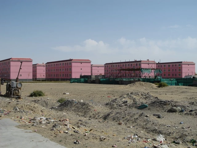 Mehrere rosa Betongebäude in staubiger Wüstenumgebung, Schuttzäune, Geröll und Müll im Vordergrund