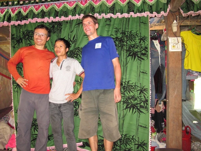 Drei Männer unterschiedlicher Größe stehen vor einem grünen Vorhang mit Bambusblattmuster, gekleidet in rotes, weißes und blaues T-Shirt