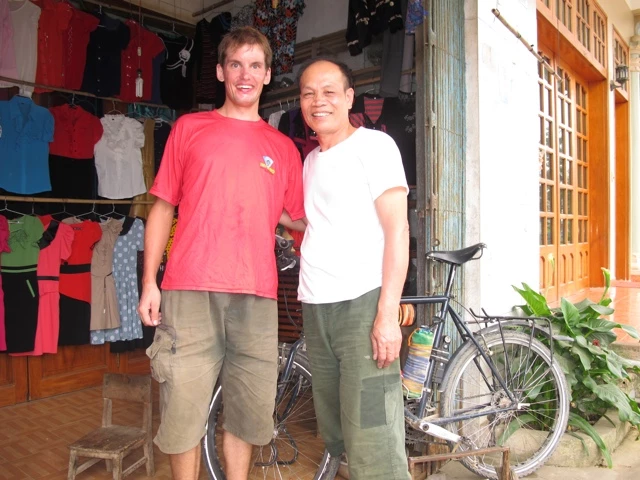 Zwei lächelnde Männer stehen in einem Bekleidungsgeschäft mit Fahrrad, bunte Shirts im Hintergrund