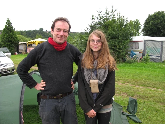 Mann und Frau in Freizeitkleidung stehen auf grüner Wiese neben grünem Auto, Wohnwagen im Hintergrund