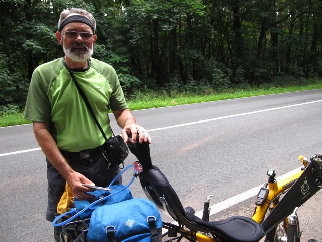 Älterer Radreisender mit grünem Shirt und Mütze steht mit Fahrrad an Waldstraße, blaue Seitentasche
