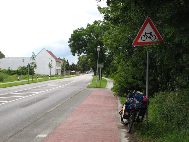 Breite Straße mit Radweg, rotem Fahrradschild und beladenem Fahrrad am Straßenrand unter Bäumen