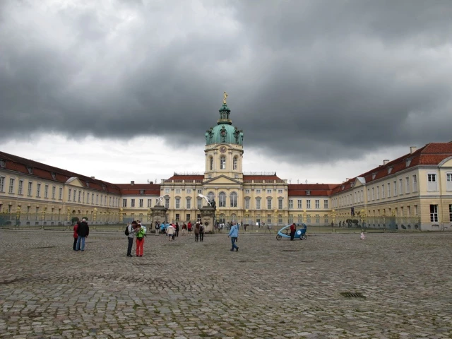 Barockes Schlossgebäude mit türkis-grüner Kuppel, graue Wolken, Kopfsteinpflaster, Touristen im Vordergrund