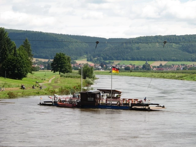 Flusslandschaft mit Fährboot, deutscher Flagge, grünen Wiesen und bewaldeten Hügeln im Hintergrund