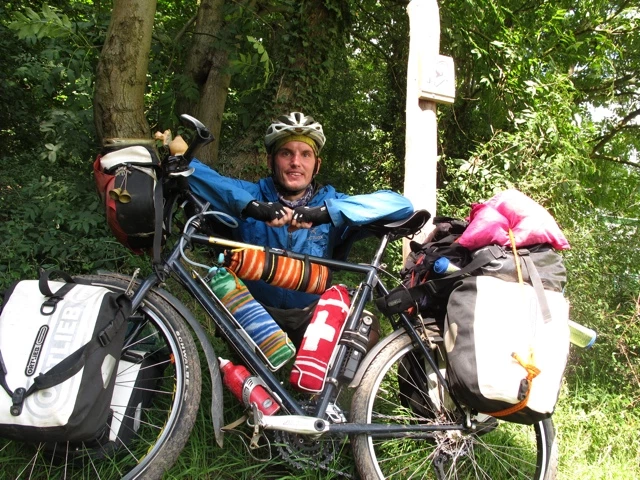 Radreisender in blauer Jacke und Helm sitzt zwischen Bäumen neben vollbepacktem Fahrrad mit Satteltaschen und Gepäck