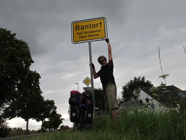 Radfahrer steht neben Ortseingangsschild von Bantorf, bewölkter Himmel, Bäume und Straße im Hintergrund