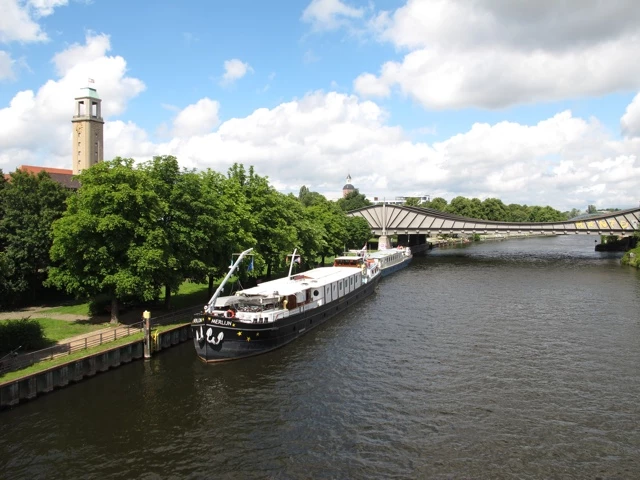 Graues Schiff an Anlegestelle neben modernem Betonbau, grüne Bäume, Turm im Hintergrund, bewölkter Himmel
