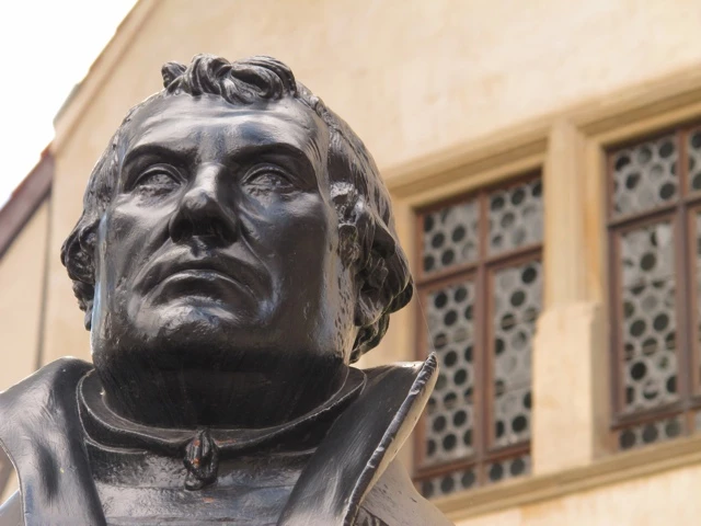 Nahaufnahme einer dunklen Bronzebüste eines Mannes mit nach oben gerichtetem Blick vor einer beigen Hauswand