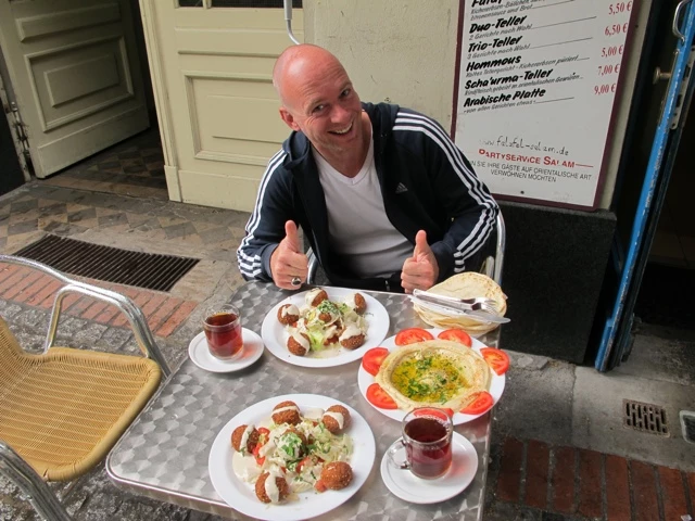 Bald gedeckter Tisch mit zwei Tellern Falafel, Salat, Tomaten und zwei Gläsern Tee. Ein lächelnder Mann im Adidas-Shirt gibt Daumen hoch