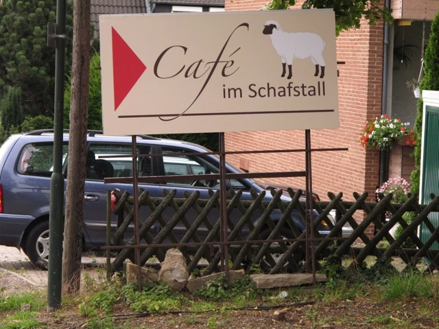 Schild für 'Café im Schafstall' mit blauem Auto, Holzzaun und Backsteingebäude im Hintergrund