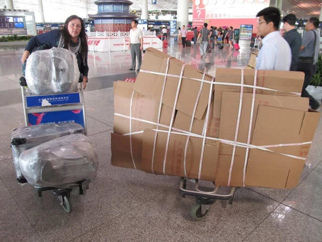 Gepäckwagen mit großen Kartons und Koffern in Flughafenhalle mit Menschen im Hintergrund