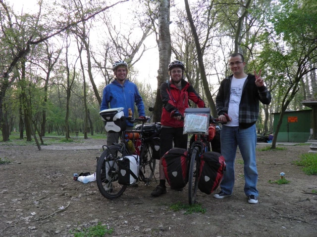 Drei Männer mit beladenen Fahrrädern stehen in einem Waldgebiet mit kahlen und belaubten Bäumen