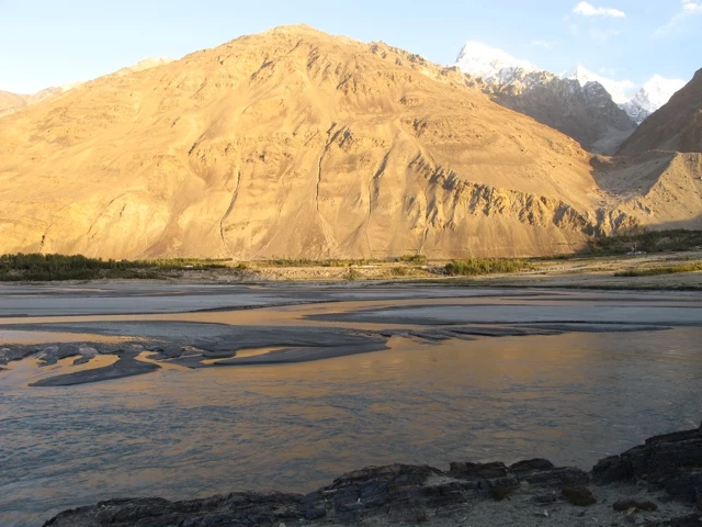 Goldgelbe Bergflanke mit steilen Felswänden, davor breites Flussbett mit Kiesbänken und Geröll in der Abendsonne